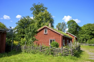 Red wooden house with grass-covered roof surrounded by trees and wooden fence under clear blue sky,