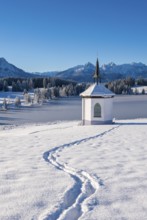 A small chapel in a snowy landscape with mountains in the background and a clear blue sky,