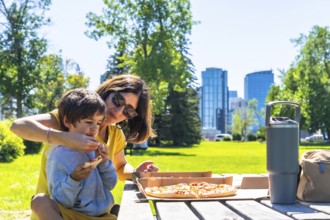 Mother helps her son eat a slice of pizza at a picnic table in a calgary park, enjoying quality