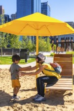 Mother and son enjoying a sunny day, playing together in a sandy urban park under a vibrant yellow
