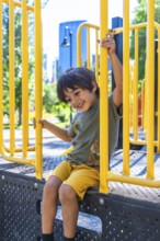 Young boy with a joyful expression sits on a playground structure, holding onto yellow bars and