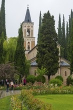 A tall church tower surrounded by blooming gardens and tall trees on a cloudy day, Bodbe monastery,