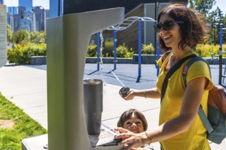 Mother and her son are filling a reusable water bottle at a public drinking fountain in a park in
