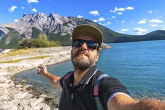 Happy tourist taking a selfie with outstretched arm by turquoise lake minnewanka, surrounded by the