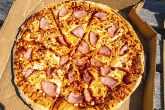 Sliced pizza with ham and melted cheese inside a cardboard box is waiting to be eaten outdoors,