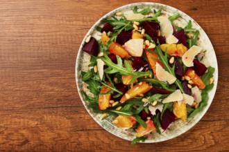 A bowl of salad contains beetroot, orange slices, pine nuts, and leaves. It is placed on a wooden