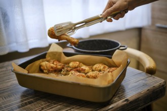 A hand uses tongs to pick up a roasted chicken drumstick from a tray in a kitchen. The tray sits on