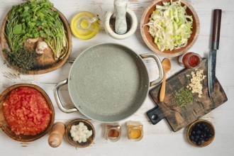 Various food items and cooking tools are arranged on a wooden table. Herbs, vegetables, and spices