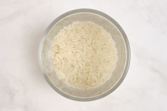 A clear bowl contains uncooked white rice. The rice is fluffy and dry, waiting to be cooked. The