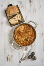 A pot with rabbit stew with vegetables and herbs on a wooden surface. Slices of bread lay on a
