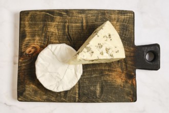 Two types of cheese on a wooden board. One cheese is a wedge with herbs. The other cheese is round