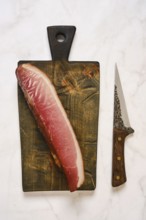 Cured parma ham on a wooden cutting board. A sharp knife is placed next to it. This scene is set in