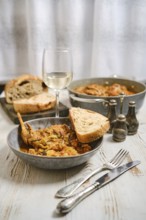 A dinner table is prepared with a bowl of food, a glass of white wine, and slices of bread. Other