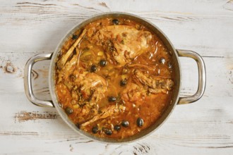 Rabbit legs are cooking in a pot filled with a sauce and olives. The pot sits on a wooden table.