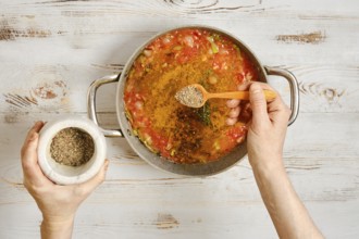 A person adds pepper to a pot of vegetables and tomatoes. The hands hold a wooden spoon and a