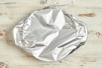 A roasting pot is covered with aluminum foil placed on a wooden kitchen table. The foil prevents