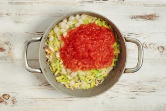 On a wooden surface, a pan holds chopped leek and pearl onions. A layer of crushed tomato sits on