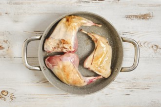 Three raw rabbit legs are browning in a metal pot on a wooden surface. The scene shows the