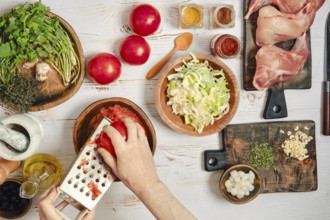 A person is grating tomatoes and chopping vegetables. Fresh herbs, spices, and rabbit meat are laid