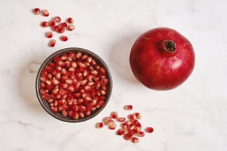 A round red pomegranate sits next to a bowl filled with bright red seeds on a white marble surface.