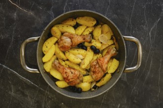 Chicken drumsticks are cooking with potato wedges and olives in a pan on a dark kitchen counter.