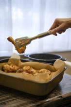 A hand lifts a piece of grilled chicken drumstick from a tray. The scene shows a cozy kitchen setup