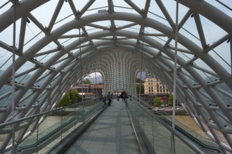 View through the modern glass bridge across a river with urban surroundings, Peace Bridge, Tbilisi,