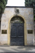 Decorated iron gate in a stone wall with plaques and surrounded by trees, entrance to the Georgian