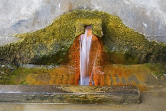 Rust-brown water flows from a spring in a concrete wall surrounded by eroded mineral deposits,