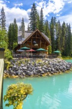 Wooden lodge with patio furniture and umbrellas reflecting in turquoise water on the shore of