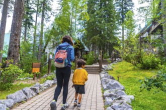 Tourists walking on a paved path approaching emerald lake lodge, surrounded by lush forest and