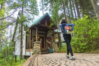 Female tourist with backpack walking up cobblestone path to a wooden cabin surrounded by lush green