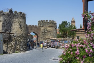 An old city gate with fortress wall and tower, animated by tourists, flanked by colorful stalls and