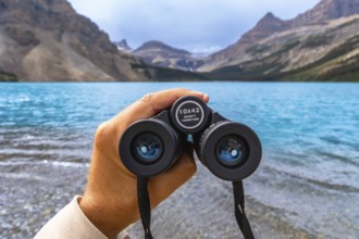 Hand holding binoculars overlooking the turquoise waters of bow lake with the canadian rockies in