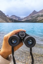 Hand holding binoculars, focusing on turquoise waters of bow lake surrounded by majestic mountains