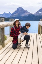 Mother happily holds her son on a wooden bridge, enjoying the stunning turquoise waters of bow lake