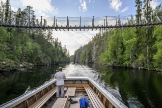 A boat sails on a calm lake under a suspension bridge between wooded banks, Julmy Ölkky, Hossq