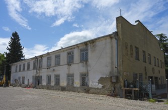 Old, dilapidated building with lattice windows under cloudy sky, excavator in the foreground,