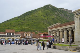 People gather in a square in front of historic buildings and a green mountain backdrop, Mtskheta