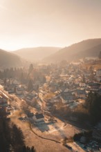 Aerial view of a snowy village in a valley at sunset with surrounding forests and hills,