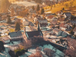 Village in winter with a central church surrounded by houses, hills and soft, warm light,