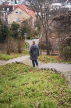 A woman in a cloak walks down an overgrown staircase surrounded by trees and houses, Herrenberg,