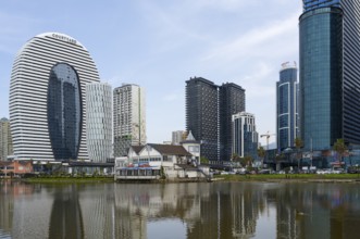 Modern high-rise buildings with glass facades are reflected in a calm lake under a cloudy sky,