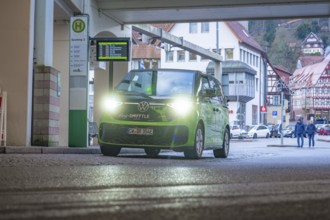 Green shuttle at a covered stop, surrounded by half-timbered houses, Deer E-Mobility Shuttle