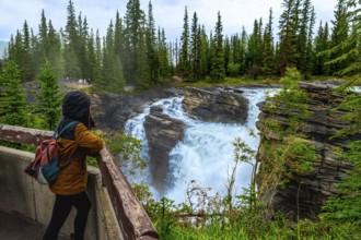 Tourist in a hooded jacket and backpack leaning on a wooden railing, admiring the powerful