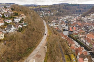 A train moves through a picturesque town, which is located along a hill and near a riverbank,
