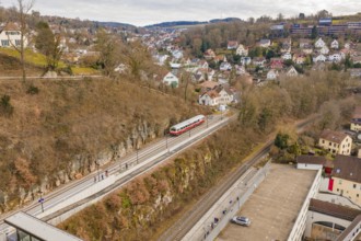 A train moves through an urban environment with hills and houses, surrounded by winter vegetation