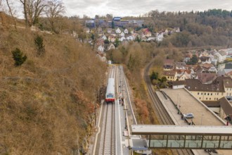 A train travels through a hilly urban landscape with wooded slopes and isolated houses under cloudy