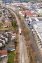 Train travels through a suburb with residential buildings and industrial buildings, surrounded by