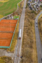 A train travels along railroad tracks past tennis courts, next to a busy road, historic event,